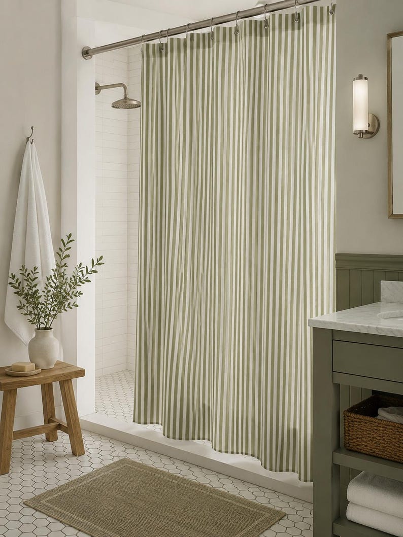 May include: A shower curtain featuring a vertical striped pattern in alternating shades of sage green and white. The fabric hangs from a silver metal rod with circular rings, creating soft folds that extend from the top to the bottom edge.