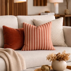 Puede incluir: Un conjunto de tres cojines cuadrados sobre un sofá beige claro. Un cojín es de terciopelo liso color óxido, otro presenta rayas verticales en color óxido y blanco, y el tercero es de tela beige claro con un sutil patrón geométrico de diamantes.