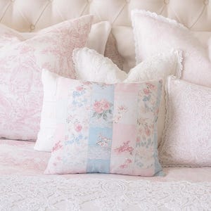 May include: A collection of decorative pillows in soft pastel colors. The central pillow features a patchwork design with floral patterns in pink, blue, and white. Other pillows have lace and floral prints, creating a romantic, vintage aesthetic.