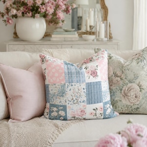 May include: A square throw pillow featuring a patchwork design of alternating squares in shades of dusty blue, soft pink, and white. The pattern includes floral motifs, small-scale gingham checks, and solid colours, creating a multi-textured appearance.