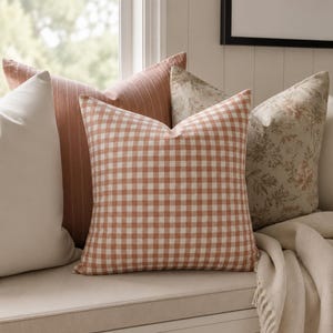May include: A collection of four square throw pillows featuring a variety of patterns including a solid cream, a terracotta striped fabric, a terracotta and white gingham check, and a beige pillow with a detailed floral botanical print.