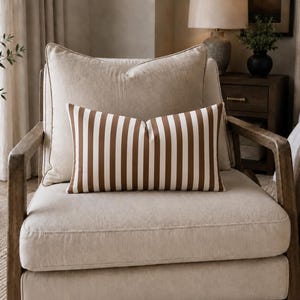Op de afbeelding: Een rechthoekig lendenkussen met een verticaal gestreept patroon in bruin en wit. Het kussen is geplaatst op een beige gestoffeerde fauteuil met houten armleuningen, voor een groter vierkant beige kussen.