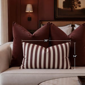 May include: A rectangular throw pillow featuring vertical stripes in dark maroon and off-white. The pillow is positioned in front of two solid dark maroon square pillows. Text on the image indicates dimensions of 20 inches in width and 14 inches in height.