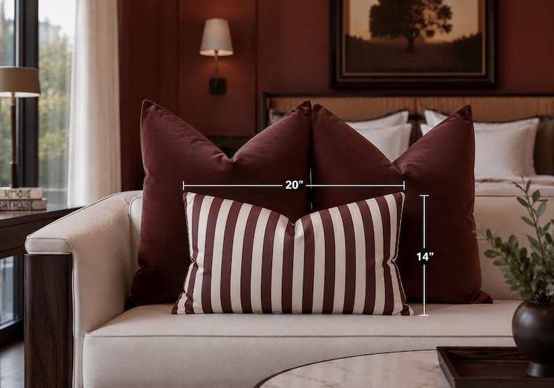 May include: A rectangular throw pillow featuring vertical stripes in dark maroon and off-white. The pillow is positioned in front of two solid dark maroon square pillows. Text on the image indicates dimensions of 20 inches in width and 14 inches in height.