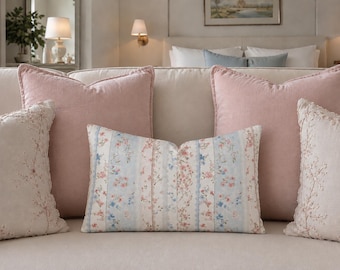May include: A collection of five throw pillows arranged on a sofa. Two large square pillows are solid dusty pink. Two square pillows are off-white with delicate floral embroidery. One central rectangular pillow features vertical stripes of floral patterns.