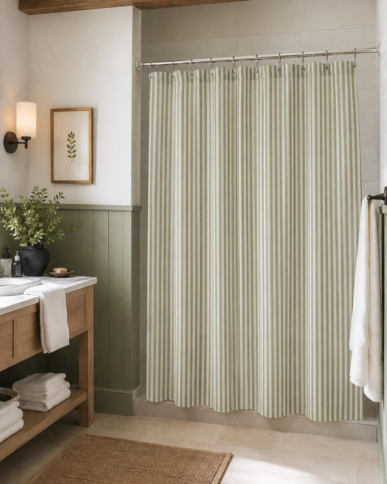 May include: A shower curtain featuring a vertical striped pattern in alternating shades of sage green and white. The fabric hangs from a silver metal rod with matching circular rings, creating uniform folds across the length of the curtain.