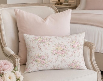 May include: A rectangular throw pillow featuring a floral pattern with pink and yellow roses on a white background, positioned in front of a solid light pink square throw pillow on a light-colored upholstered armchair with a wooden frame.