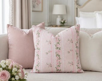 May include: A square throw pillow featuring a light pink background with vertical stripes of white lace patterns and small pink rose floral motifs. The pillow is positioned on a sofa between a solid light pink pillow and a solid cream pillow.