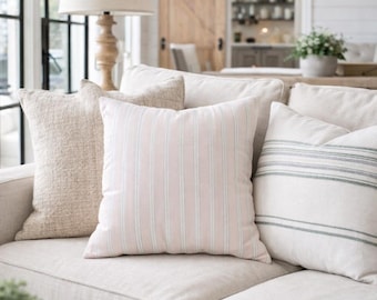 May include: A square throw pillow featuring a light pink and white vertical striped pattern. The pillow is positioned on a light beige sofa alongside other textured throw pillows in neutral tones, creating a coordinated arrangement of soft home textiles.