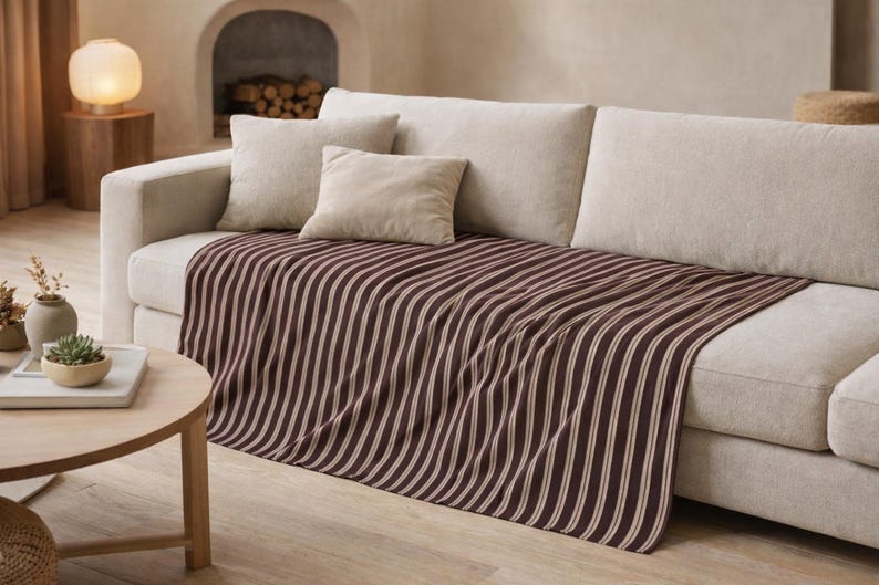 Puede incluir: Sof&aacute; crema con cojines beige y una manta a rayas marr&oacute;n y blanca. Una mesa de centro redonda de madera con una planta y una l&aacute;mpara est&aacute;n delante. La habitaci&oacute;n tiene una combinaci&oacute;n de colores neutros.