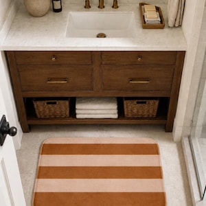 May include: A rectangular bathroom floor mat featuring wide, horizontal stripes in alternating shades of terracotta orange and soft beige. The mat has rounded corners and a plush, uniform texture, positioned on a light-colored tiled floor.