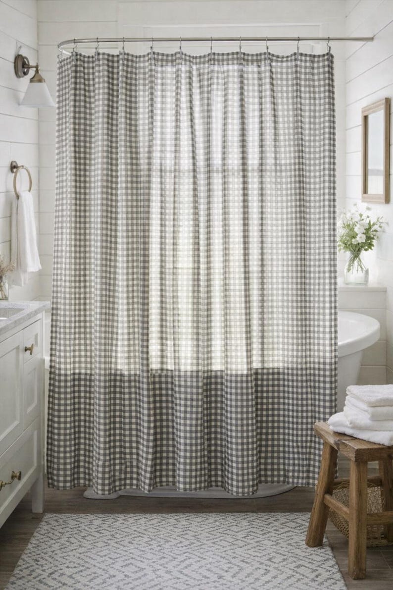 May include: A gray and white gingham shower curtain hangs in a bathroom. The curtain is hung from a curved rod and has a darker gray border at the bottom. The bathroom has white walls and a white vanity.