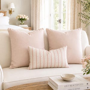 May include: Two large square light pink throw pillows and one smaller rectangular lumbar pillow with vertical white and pink stripes. The pillows are arranged on a cream-coloured sofa, providing a soft, neutral aesthetic for a living space.