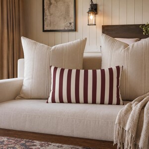 May include: Two large square beige throw pillows with subtle vertical stitching and one rectangular lumbar pillow featuring bold, vertical dark red and white stripes. The pillows are arranged on a light beige sofa with a textured throw blanket.