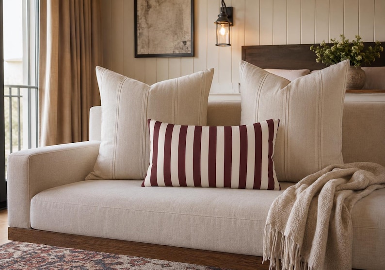 May include: Two large square beige throw pillows with subtle vertical stitching and one rectangular lumbar pillow featuring bold, vertical dark red and white stripes. The pillows are arranged on a light beige sofa with a textured throw blanket.