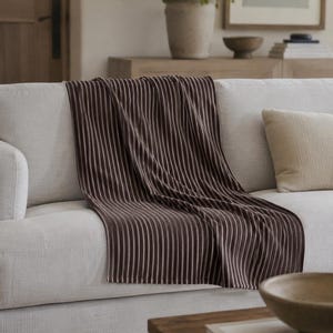 Puede incluir: Una manta a rayas marrones y blancas sobre un sof&aacute; de color claro. Las rayas verticales de la manta crean una textura visual. Un coj&iacute;n beige y una mesa de centro de madera con un cuenco y un libro est&aacute;n en primer plano.