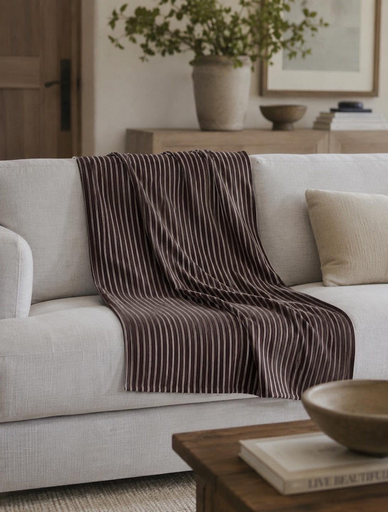 Puede incluir: Una manta a rayas marrones y blancas sobre un sof&aacute; de color claro. Las rayas verticales de la manta crean una textura visual. Un coj&iacute;n beige y una mesa de centro de madera con un cuenco y un libro est&aacute;n en primer plano.