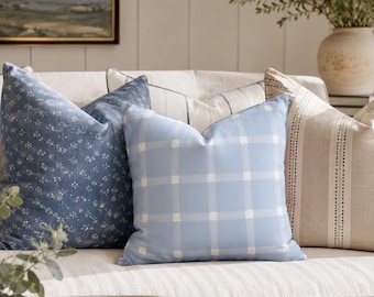 May include: A collection of three square throw cushions arranged on a sofa. The left cushion is dark blue with a small white floral pattern. The centre cushion is light blue with a white grid check pattern. The right cushion is beige with vertical stripes.