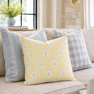 May include: A square throw pillow featuring a yellow background with a repeating pattern of white oval frames containing small blue flowers and green stems, flanked by a blue striped pillow and a blue and white gingham checkered pillow.
