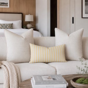 Peut inclure: Un canapé crème avec trois coussins et un coussin lombaire à rayures jaunes. Un plaid en tricot beige est drapé sur le bras. Une table basse en bois contient des livres et un petit bol en bois avec de la verdure.