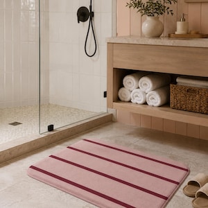 Peut inclure: Un tapis de bain rectangulaire présentant une base rose poudré douce avec quatre rayures horizontales distinctes et régulièrement espacées d'un rouge bordeaux profond. Le tapis a des coins arrondis et une texture lisse et uniforme.