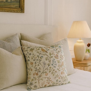 May include: A decorative square pillow with a botanical design featuring a bird, flowers, and vines in shades of green, blue, and orange on a cream background. The pillow is part of a set of pillows on a bed with a cream-colored headboard.