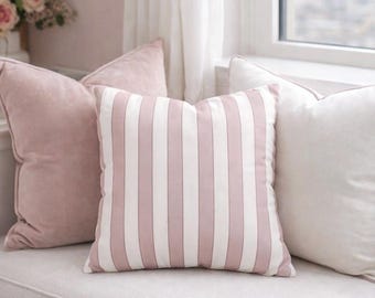 May include: A collection of decorative throw pillows in shades of pink and white. One pillow features vertical stripes, while the others are solid. The pillows are arranged on a light-colored bench, creating a soft and inviting aesthetic.