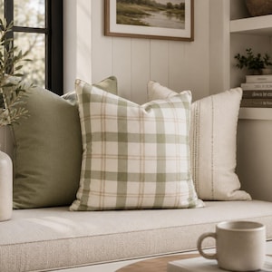Peut inclure: Trois coussins carrés disposés en rangée. Le coussin de gauche est vert sauge uni. Le coussin central présente un motif à carreaux vert sauge et crème. Le coussin de droite est crème avec des rayures verticales texturées et un tissage subtil.