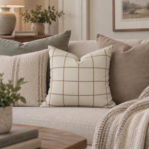 May include: A square throw pillow featuring a cream-colored fabric with a thin, brown grid pattern. The pillow is positioned centrally among other decorative cushions on a sofa, showcasing a clean, geometric design with visible stitching.