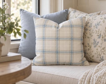 Op de afbeelding: Een vierkant sierkussen met een lichtcrème basis en een blauw en beige ruitpatroon. Het kussen staat in het midden, geflankeerd door een effen lichtblauw kussen aan de linkerkant en een kussen met bloemenpatroon aan de rechterkant.
