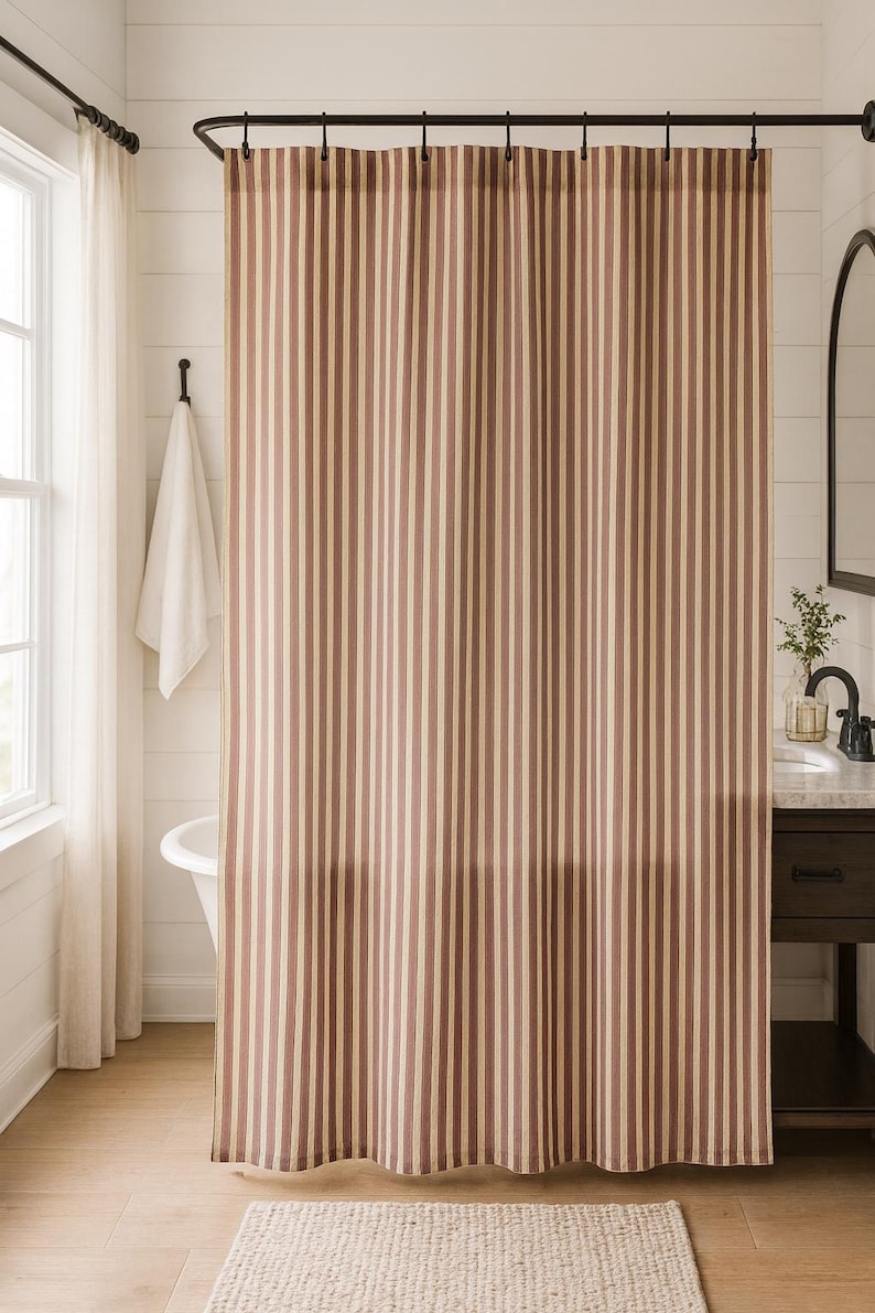 May include: A shower curtain featuring a vertical striped pattern in alternating shades of muted reddish-brown and cream. The curtain hangs from a black metal rod with integrated rings, displaying a consistent pleated drape across its full width.