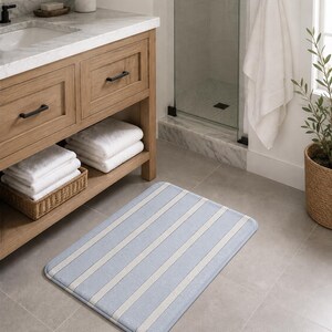 May include: A rectangular bathroom rug featuring a pattern of light blue and off-white vertical stripes. The rug has rounded corners and a soft, plush texture, positioned on a gray tiled floor in front of a wooden vanity cabinet.