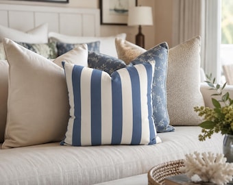 May include: A square throw pillow featuring vertical navy blue and off-white stripes. The pillow is positioned in the center of a collection of various decorative cushions, including solid beige, textured cream, and blue floral patterned pillows.