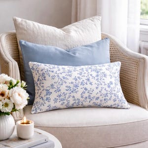 Peut inclure: Un fauteuil crème avec trois coussins aux tons de blanc et de bleu. Le coussin avant présente un fond blanc avec un motif floral bleu. Une petite table avec une bougie et des fleurs est devant la chaise, créant une ambiance chaleureuse.