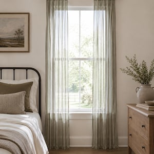 Puede incluir: Interior de dormitorio con cortinas transparentes a rayas verde oliva. Se ve una cama con ropa de cama blanca y cojines, junto con una cómoda de madera y una pintura de paisaje enmarcada. Una alfombra tejida está en el suelo.