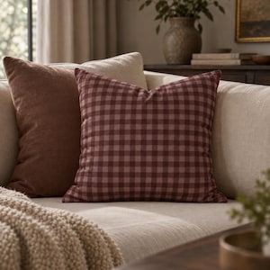 May include: A square throw pillow featuring a dark red and light pink gingham check pattern. The pillow is positioned on a sofa next to a solid brown throw pillow and a textured cream-colored throw blanket, creating a layered arrangement.