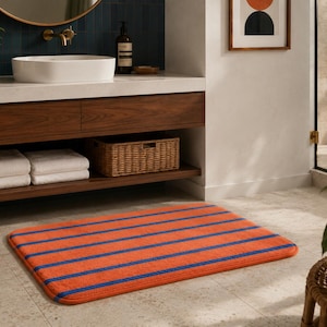 Peut inclure: Un tapis de bain rectangulaire présentant une base orange vif avec des rayures horizontales bleu marine. Le tapis possède des coins arrondis et une surface douce et texturée, posé sur un sol carrelé clair devant un meuble vasque en bois.