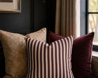 Può includere: Una collezione di cuscini quadrati composta da un cuscino centrale a righe verticali bordeaux e crema, affiancato da un cuscino in velluto bordeaux scuro a tinta unita e da un cuscino color cuoio con un delicato motivo floreale bianco.
