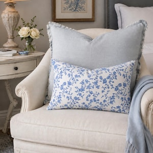 May include: A square light blue pillow with frayed edges and a rectangular white pillow featuring a blue floral pattern. Both pillows are placed on a cream-colored armchair, accompanied by a light blue throw blanket draped over the side of the chair.