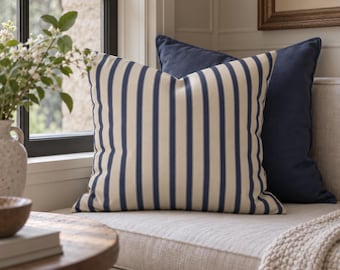 May include: A square throw pillow featuring a vertical striped pattern in navy blue and cream. The pillow is positioned in front of a solid navy blue throw pillow. Both items are placed on a light-colored upholstered bench seat.