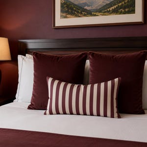May include: A bed featuring a white duvet and a dark burgundy runner, two large square burgundy velvet pillows, two white rectangular pillows with dark piping, and one rectangular lumbar pillow with vertical burgundy and cream stripes.