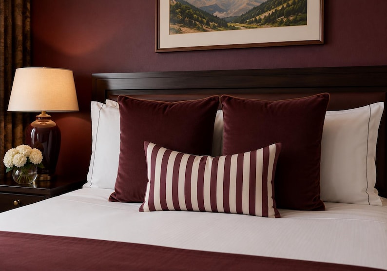 May include: A bed featuring a white duvet and a dark burgundy runner, two large square burgundy velvet pillows, two white rectangular pillows with dark piping, and one rectangular lumbar pillow with vertical burgundy and cream stripes.