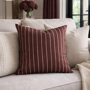 May include: A decorative throw pillow with a dark brown background and vertical white stripes. The pillow is square and rests on a light-colored sofa, alongside other pillows and a blanket. The setting appears to be a living room.