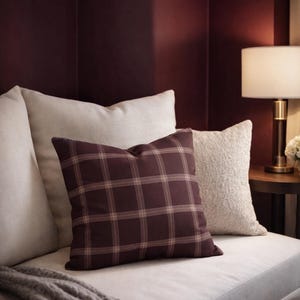 Könnte beinhalten: Ein weinrotes kariertes Zierkissen liegt auf einem weißen Sofa, neben einem strukturierten cremefarbenen Kissen. Eine graue Strickdecke ist über das Sofa drapiert. Der Hintergrund zeigt eine dunkelweinrote Wand und eine Tischlampe.