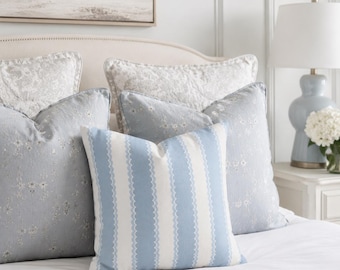 May include: A square throw pillow featuring vertical light blue and white stripes with a wavy line pattern, positioned in front of two larger square pillows with a light blue floral print and two quilted white pillows on a bed.