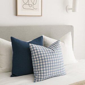 Pu&ograve; includere: Una collezione di cuscini quadrati disposti su un letto. Il set comprende due cuscini bianchi a tinta unita, un cuscino blu navy a tinta unita e un cuscino con motivo a quadretti vichy blu e bianchi, tutti appoggiati su un copriletto bianco.