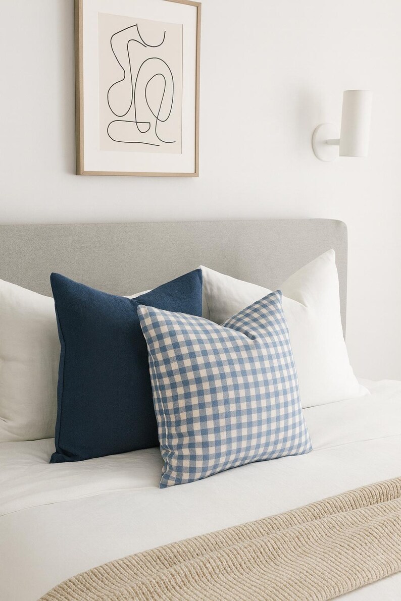 Pu&ograve; includere: Una collezione di cuscini quadrati disposti su un letto. Il set comprende due cuscini bianchi a tinta unita, un cuscino blu navy a tinta unita e un cuscino con motivo a quadretti vichy blu e bianchi, tutti appoggiati su un copriletto bianco.