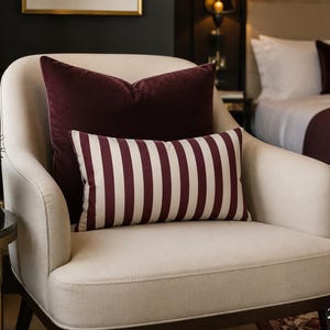 May include: A rectangular throw pillow featuring a vertical striped pattern in deep burgundy and off-white. The pillow measures 20 inches by 14 inches and is positioned in front of a solid burgundy square pillow on a cream-colored upholstered armchair.