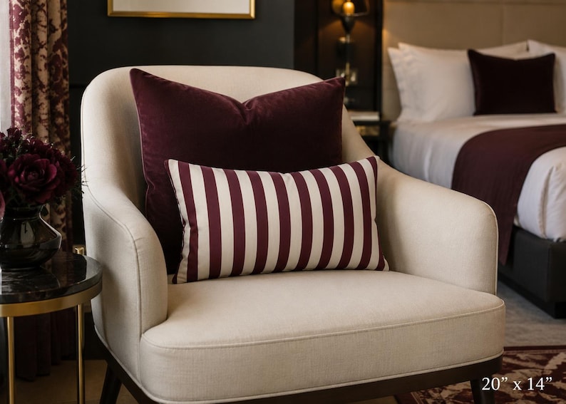May include: A rectangular throw pillow featuring a vertical striped pattern in deep burgundy and off-white. The pillow measures 20 inches by 14 inches and is positioned in front of a solid burgundy square pillow on a cream-colored upholstered armchair.