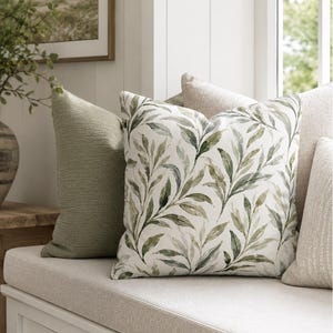 May include: A square throw pillow featuring a white background with a repeating watercolor-style pattern of green leaves. The pillow is positioned alongside a solid sage green textured pillow and a plain cream-colored pillow on a bench seat.